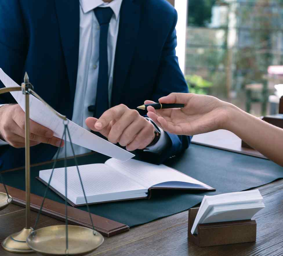 A lawyer passing a pen across a desk to a client, with brass scales of justice and an open document in the foreground