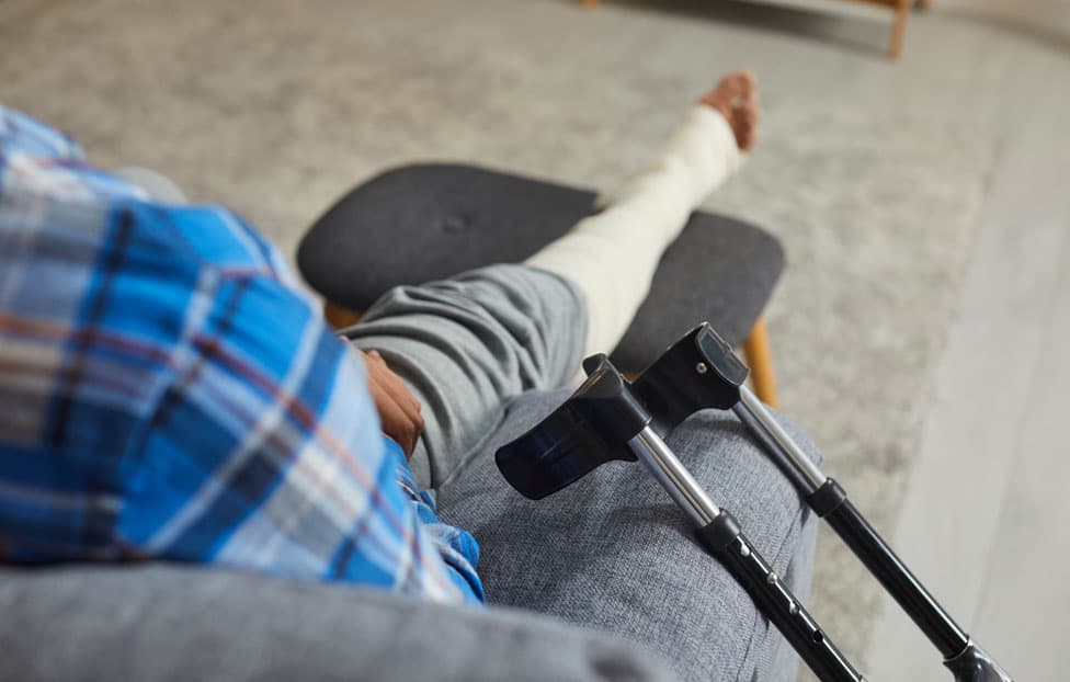 A person resting on a sofa with a cast on their lower leg and crutches propped beside them