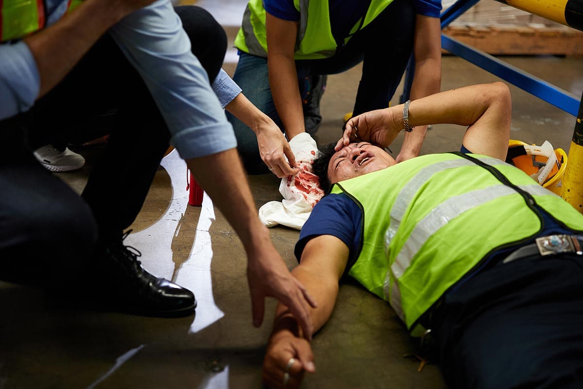 Colleagues in high-visibility vests tending to an injured warehouse worker lying on the floor